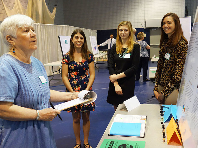 A judge reviews a poster presentation, while three female students provide additional insight