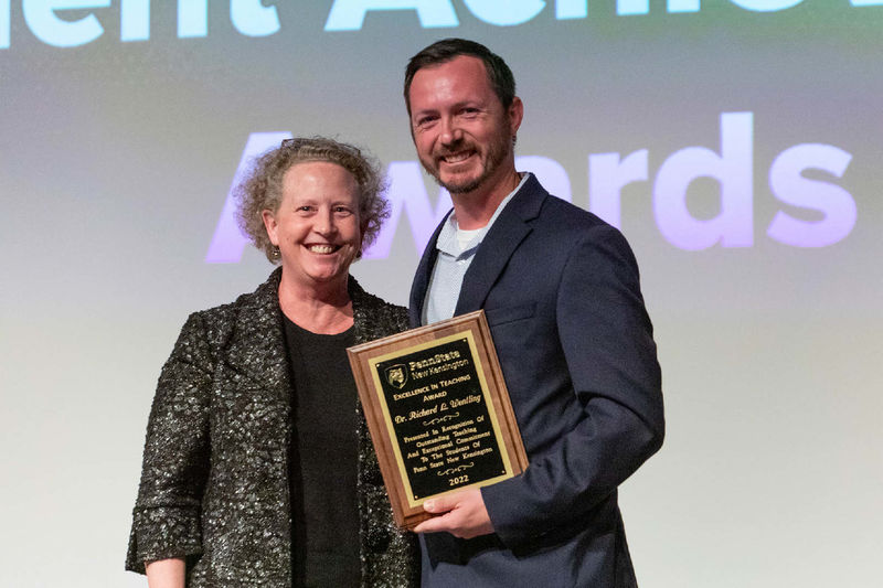 Faculty member holds plaque and stands next to Director of Academic Affairs