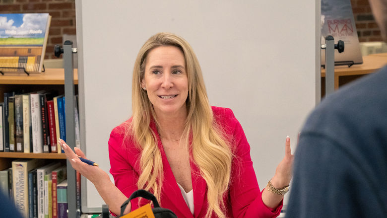 Erin Weber, dressed in a bright pink blazer, gestures speaking to students at a table in the library.