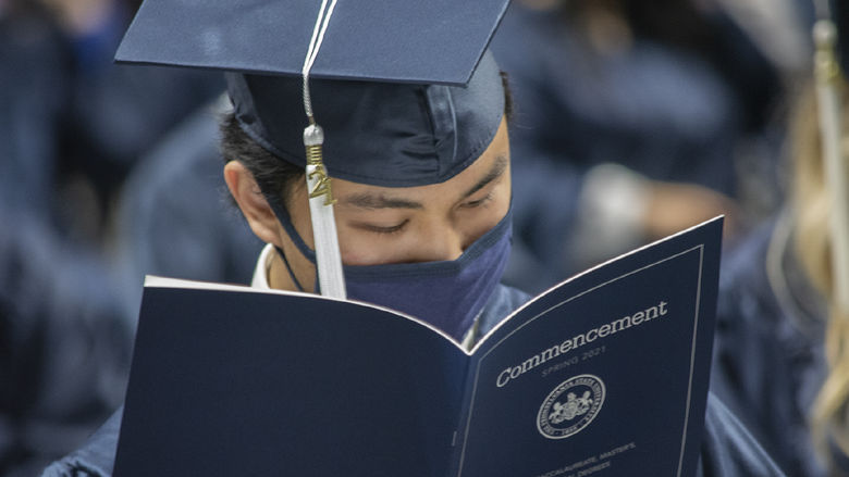 graduate in cap, gown, and face mask reading program