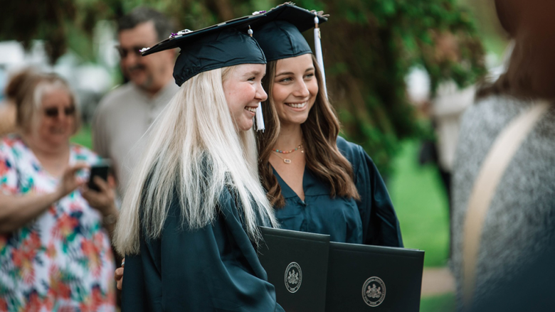 : Two graduates in caps and gowns smile while holding their diplomas during as friends and family take photos in the background.