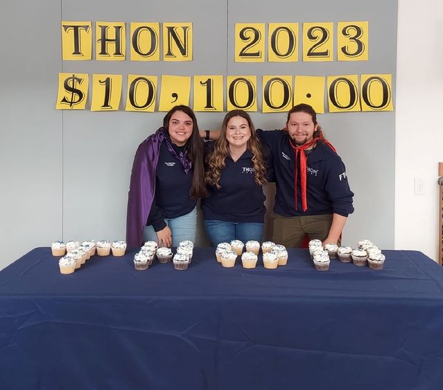 Three students stand smiling by table