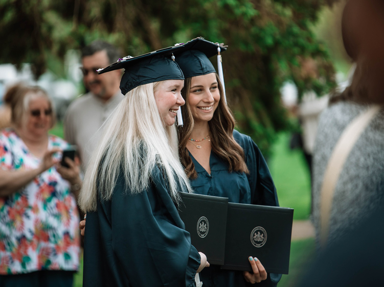 : Two graduates in caps and gowns smile while holding their diplomas during as friends and family take photos in the background.